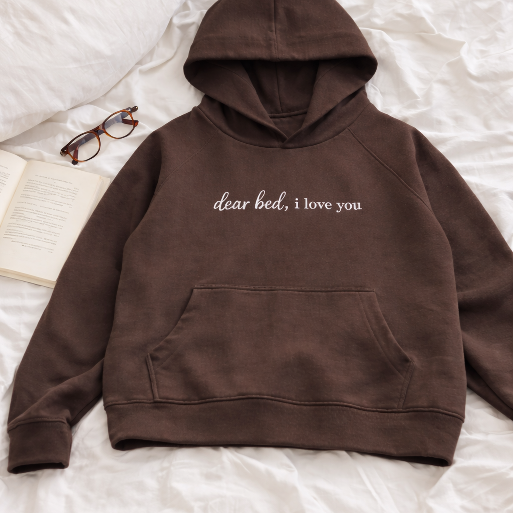 Brown hoodie with 'dear bed, i love you' text on a white bedspread.