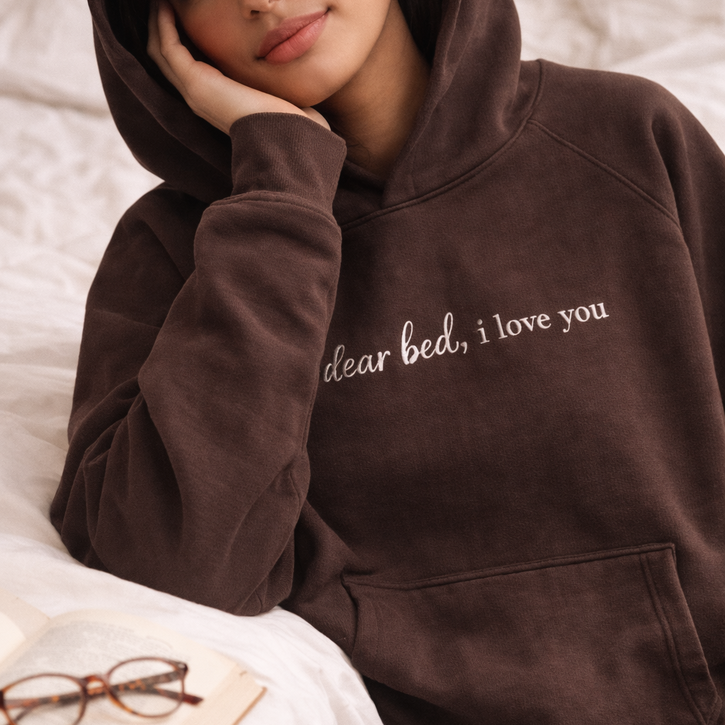 Person wearing a brown hoodie with 'dear bed, i love you' text, sitting on a bed.