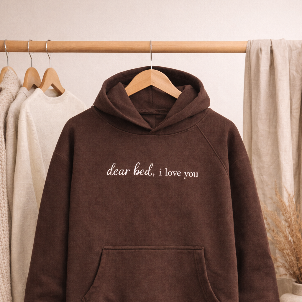 Brown hoodie with 'dear bed, i love you' text on a hanger against a neutral background